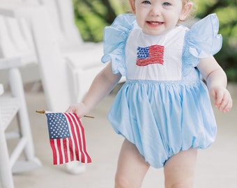 May include: A baby girl wearing a light blue dress with a red, white, and blue embroidered American flag on the front. She is holding a small American flag in her right hand and has a red bow in her hair.