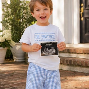May include: A white short-sleeved t-shirt featuring a blue smocked panel with the text BIG BROTHER in white, paired with light blue and white gingham shorts. The garment is shown with a small rectangular ultrasound scan held in front.