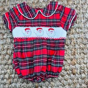 May include: A red and green plaid baby romper with a white collar and three embroidered Santa faces on the chest.