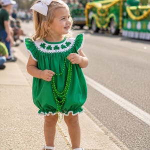 May include: A green bubble romper featuring a white yoke with embroidered green shamrocks, ruffled sleeves, and white trim. The garment is paired with a large white hair bow and a strand of green plastic beads, worn with white sandals.