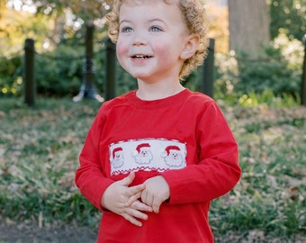 Puede incluir: Un niño pequeño con una camisa roja de manga larga con un diseño bordado blanco de tres caras de Papá Noel. El niño sonríe y mira hacia arriba.