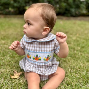 May include: A baby wearing a brown and white gingham romper with a blue-trimmed collar and sleeves. The romper features an embroidered design of colorful turkeys. The baby is sitting on green grass.