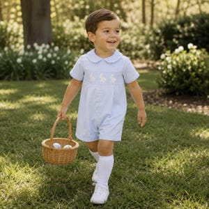 May include: A light blue short-sleeved romper featuring a Peter Pan collar and a smocked chest panel with white embroidered bunny rabbits. The garment is paired with white knee-high socks and white shoes, while holding a small woven wicker basket.