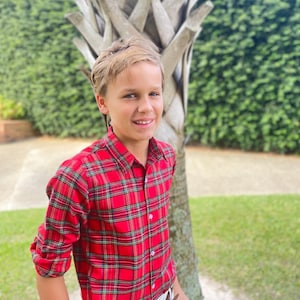 以下が含まれることがあります： 赤いチェック柄のボタンダウンシャツに茶色のベルトと白いズボンを着用した若い人。ヤシの木の前に立っています。