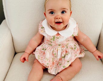 May include: A baby girl wearing a pink and white floral print romper with a white peter pan collar and a green bow headband. The baby is sitting on a white chair.