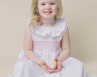 May include: A young child wearing a white dress with a pink smocked bodice and a large pink bow in their hair. The dress has a ruffled collar. The child is holding a light-colored egg. The background is a solid, light beige color.