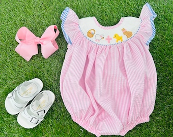 May include: A pink and white striped baby girl's bubble romper with embroidered chicks, a lamb, and a basket of eggs. The romper has blue and white ruffled sleeves.