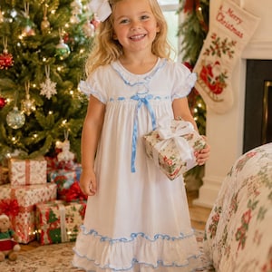 May include: A white nightgown featuring short ruffled sleeves, a round neckline with blue trim, and a blue ribbon tie at the chest. The hem is finished with two tiers of ruffles edged in blue. The garment is made of a lightweight fabric.