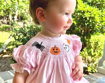 May include: A light pink dress with short, ruffled sleeves. The dress features a smocked bodice with embroidered Halloween-themed designs, including a black spider, an orange jack-o'-lantern, and a white ghost. The dress is worn by a young child.