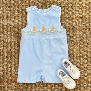 May include: A light blue baby romper with embroidered bunnies and green leaves. The romper has a white trim and buttons on the shoulders. The romper is paired with white sneakers.