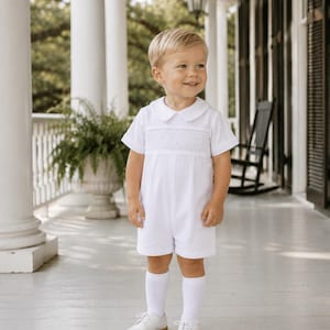 May include: A white short-sleeve romper featuring a Peter Pan collar and a smocked chest panel with intricate embroidery. The garment is paired with plain white knee-high socks and white leather shoes with a lace-up front and tan soles.