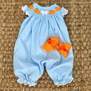Könnte beinhalten: Ein hellblaues Baby-Smocks-Kleid mit Kürbis-Stickerei und einer weißen Schleife. Das Kleid hat weiße Rüschen am Ausschnitt und an den Ärmeln. Eine passende orange Schleife ist im Lieferumfang enthalten.