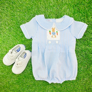 May include: A blue and white striped baby onesie with a Peter Rabbit embroidered design. The onesie has a white Peter Rabbit with a blue jacket and carrot in his hand. The onesie has a white collar with blue trim.