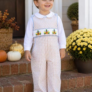 May include: A long-sleeve white shirt worn under a tan and white gingham overall jumpsuit. The jumpsuit features a smocked chest panel with embroidered colorful turkeys and blue trim, paired with a white rounded collar and white sneakers.