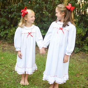 Puede incluir: Dos niñas con camisones blancos a juego con ribetes rojos y lazos en el pelo. Los camisones de manga larga tienen un dobladillo con volantes y detalles de cinta roja. Se dan la mano y están de pie sobre hierba verde.
