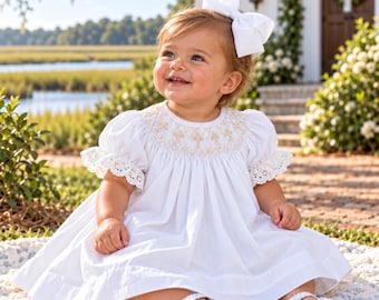 May include: A white dress featuring short puff sleeves with eyelet lace trim and a smocked bodice with gold embroidery. The dress is paired with a large white hair bow and white buckled sandals, creating a classic look for a young child.