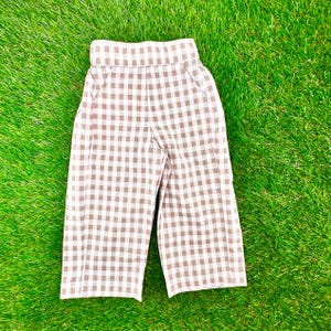 May include: A pair of brown and white checkered pants. The pants have a high waist and pockets. The fabric is a light, breathable material, perfect for warm weather. The pants are laid out on a green surface.