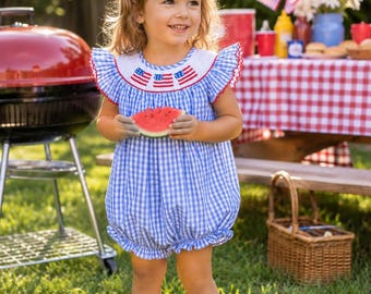 May include: A blue and white gingham bubble romper featuring a white smocked bodice with embroidered American flag motifs, ruffled sleeves with red trim, and a red hair bow. The outfit is paired with red sneakers and a slice of watermelon.