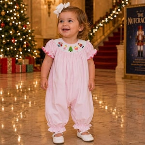 May include: A light pink bubble romper featuring short puffed sleeves with ruffled edges and a white round collar. The collar is embroidered with small colorful nutcracker figures and a green Christmas tree. A white bow is attached to the hair.