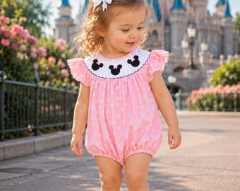 May include: A light pink bubble romper featuring a white polka dot pattern and a white smocked yoke decorated with black Minnie Mouse head silhouettes. The garment has ruffled sleeves and a small white bow hair accessory is worn on the head.