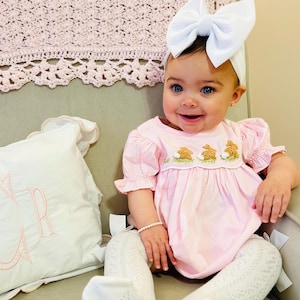 May include: A baby girl wearing a pink dress with embroidered bunnies and a white bow headband. She is sitting in a chair with a white pillow.