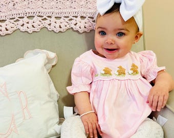 May include: A baby girl wearing a pink dress with embroidered bunnies and a white bow headband. She is sitting in a chair with a white pillow.
