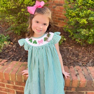 May include: A young girl wearing a green and white gingham dress with embroidered shamrocks and a pink bow. The dress has a ruffled collar and short sleeves.