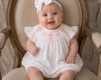 May include: A white bubble romper featuring pink smocked detailing with cross motifs across the chest, flutter sleeves, and lace trim. The outfit is paired with a white headband featuring a large bow and white leather buckled shoes.