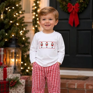 May include: A white long-sleeve shirt featuring a central smocked panel with three embroidered nutcracker figures, paired with red and white gingham trousers. The shirt has a crew neckline and the trousers have a classic straight-leg cut.