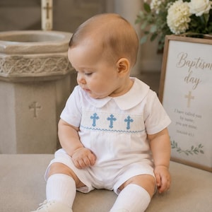 Könnte beinhalten: Ein weißer Kurzarm-Strampler mit Bubikragen und einem gesmokten Brustbereich, der mit blauen, gestickten Kreuzmotiven verziert ist. Das Outfit wird durch weiße Rippsocken und weiße Lederschuhe mit Schnürsenkeln vervollständigt.