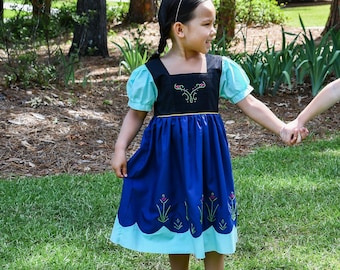 May include: A young girl wearing a blue and black dress with a white trim and floral embroidery. She is wearing a tiara and holding hands with another person.