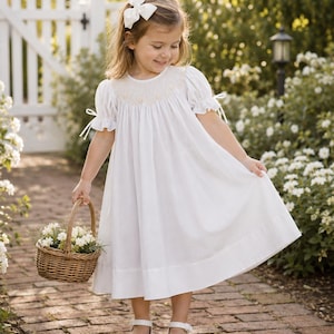 May include: A white dress with short puff sleeves, a smocked bodice featuring subtle embroidery, and a wide hem. The dress includes small ribbon ties on the sleeves. A white hair bow is worn, and a small woven basket with white flowers is held.