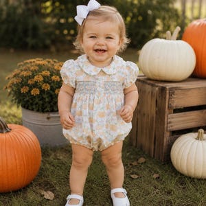 May include: A short-sleeved romper featuring a light-coloured floral and pumpkin print with a smocked bodice and a rounded collar. The garment is paired with a white hair bow and white buckled shoes, creating a coordinated outfit for a young child.