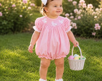 May include: A light pink bubble romper featuring white smocking with embroidered bunny details, puff sleeves with white lace trim, and a large white hair bow. The outfit is paired with white buckled shoes and a small woven basket filled with colorful eggs.