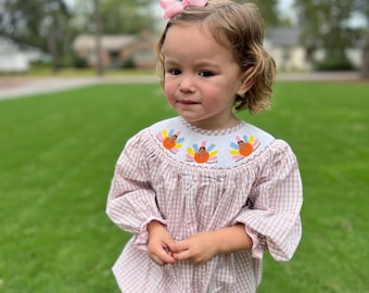 May include: A young child wearing a pink and white gingham dress with embroidered turkeys on the collar. The dress has puffed sleeves and a gathered skirt. The child is standing on a grassy lawn.