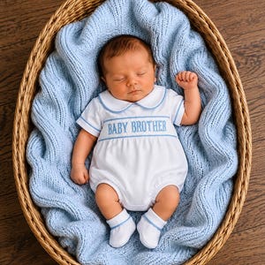 Könnte beinhalten: Ein Neugeborenes schläft in einem Weidenkorb auf einer hellblauen Strickdecke. Das Kind trägt einen weißen kurzärmeligen Strampler mit Bubikragen und blauer Umrandung, der mit dem Text BABY BROTHER in blauem Garn bestickt ist.