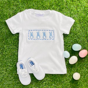 May include: White short-sleeved shirt with blue bunny embroidery and a blue and white trim. The shirt is paired with white sneakers with blue accents.