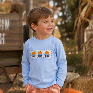 May include: A light blue long-sleeve shirt featuring a rectangular smocked panel across the chest. The panel displays three orange pumpkins in blue wagons, bordered by a blue and white checkered pattern on a white background.