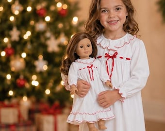 May include: A white long-sleeved nightgown featuring a ruffled collar, cuffs, and hem with red trim, and a red ribbon tie at the neckline. The garment is worn by a doll and a person, both dressed in matching white nightgowns with red accents.