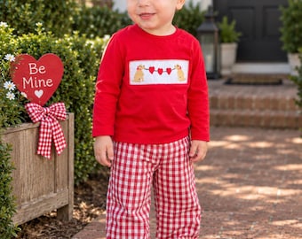 Puede incluir: Una camiseta roja de manga larga con un panel blanco fruncido que muestra dos perros dorados sosteniendo una cuerda de tres corazones rojos, combinada con pantalones de cuadros vichy rojos y blancos y zapatillas blancas.