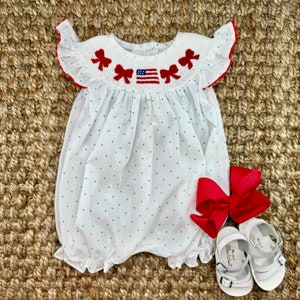 May include: White baby smocked dress with blue polka dots and red embroidered bows around the neckline. The dress features a small embroidered American flag on the front. The dress is paired with a red bow and white sandals.