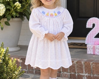 May include: A white long-sleeve dress with a small pink polka dot pattern, featuring a smocked neckline with embroidered colorful birthday cake motifs, a scalloped hem, a lavender hair bow, white knee-high socks, and white leather shoes.