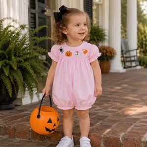 May include: A light pink bubble romper featuring short ruffled sleeves and a smocked chest with embroidered Halloween motifs including a black spider, an orange pumpkin, a white ghost, and a piece of candy corn, paired with white trainers.