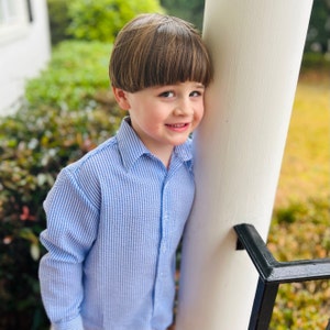 May include: A young child wearing a light blue and white striped button-down shirt. The shirt has long sleeves and a classic collar. The child is smiling and leaning against a white column.