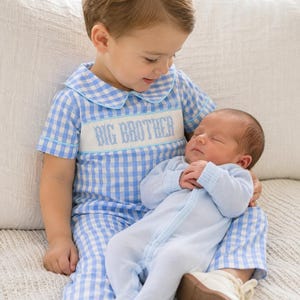 May include: A blue and white gingham short-sleeved romper with a Peter Pan collar and a white smocked chest panel featuring the embroidered text BIG BROTHER in blue, worn with a light blue long-sleeved footed sleepsuit with a front snap closure.