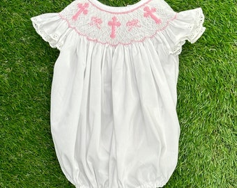 May include: White baby bubble romper with pink cross embroidery on the yoke. The romper features ruffled sleeves with eyelet detailing. The garment is made of a lightweight fabric and has a gathered bottom.