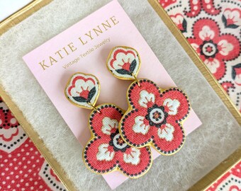 May include: Floral drop earrings with red and white patterns, presented in a gold-trimmed box. The earrings are from Katie Lynne Vintage Textile Jewelry. The earrings have a gold border and a flower design.