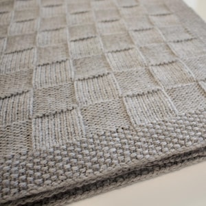 Może przedstawiać: Szary dzianinowy koc z kratką. Koc jest złożony na pół, a krawędzie są widoczne.