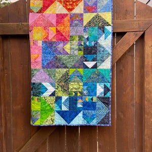 May include: A colorful patchwork quilt with a variety of geometric shapes and patterns in shades of pink, purple, blue, green, and yellow. The quilt is hanging on a wooden fence.