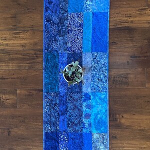 Op de afbeelding: Een blauwe en witte patchwork tafelrunner met verschillende bloemen- en geometrische patronen. De runner is gemaakt van stof en heeft een gestikte textuur.